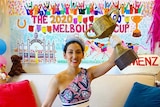 Wearing a bright dress and hat, Angela Menz holds a replica of the Melbourne Cup in a decorated lounge room.