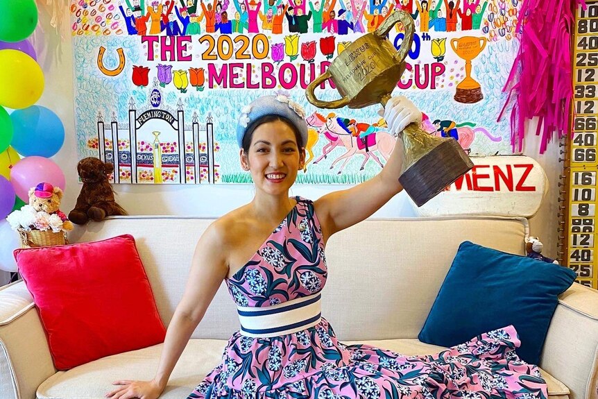 Wearing a bright dress and hat, Angela Menz holds a replica of the Melbourne Cup in a decorated lounge room.