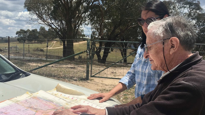 An old man and a young woman looking at a map spread on the bonnet of a car