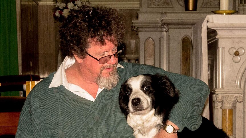 A priest, in ordinary clothes, with his arm around his borde collie dog in front of a marble altar in a Catholic Church
