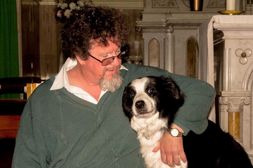 A priest, in ordinary clothes, with his arm around his borde collie dog in front of a marble altar in a Catholic Church