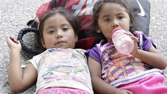 Two girls travelling on the migrant caravan rest their head on a backpack.