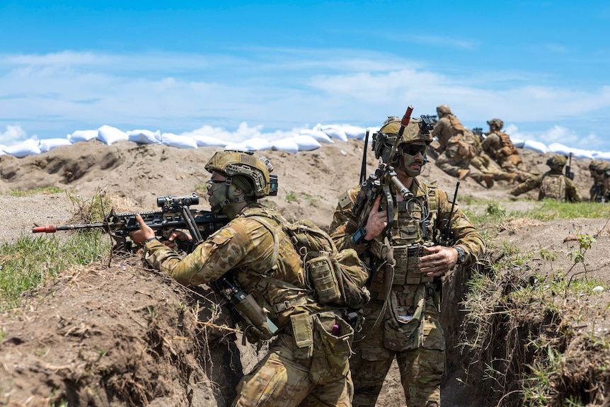 Soldiers wearing camouflage and carrying weapons while standing in a ditch with others behind them on a hill and blue sky above.