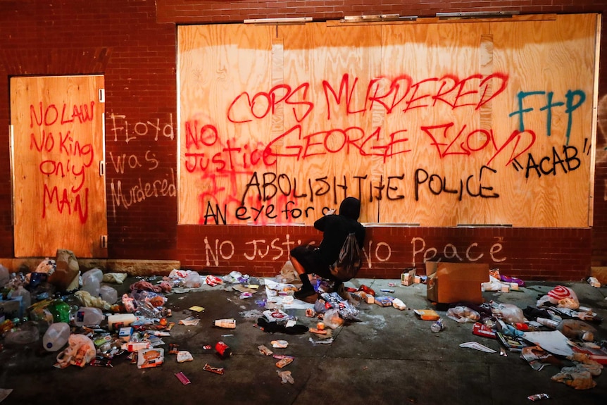 Various scrawls of graffiti are seen on a brick and wood wall, with rubbish on the ground below.