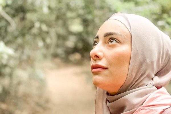 A close up photo of Aisha, who wears a hijab, looking up to the side. Trees in the backdrop.
