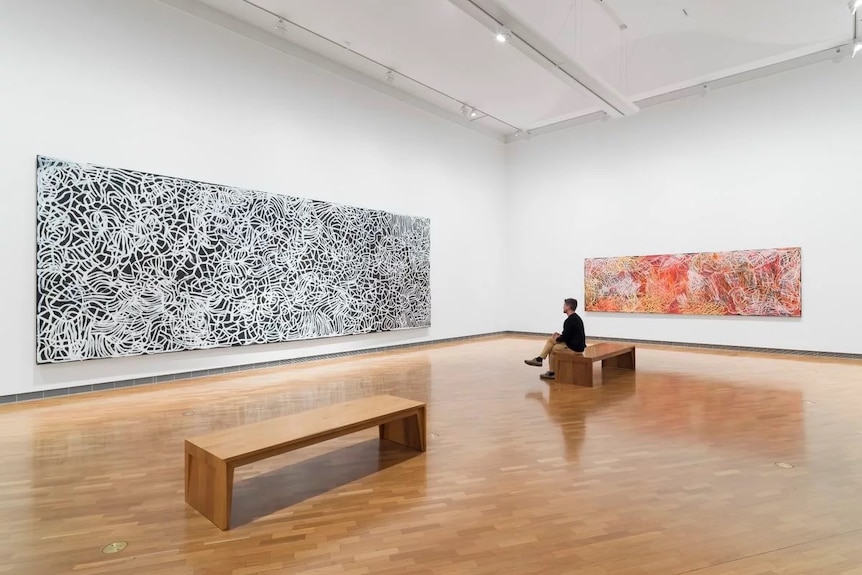 A figure sits in the gallery on a bench viewing a large artwork