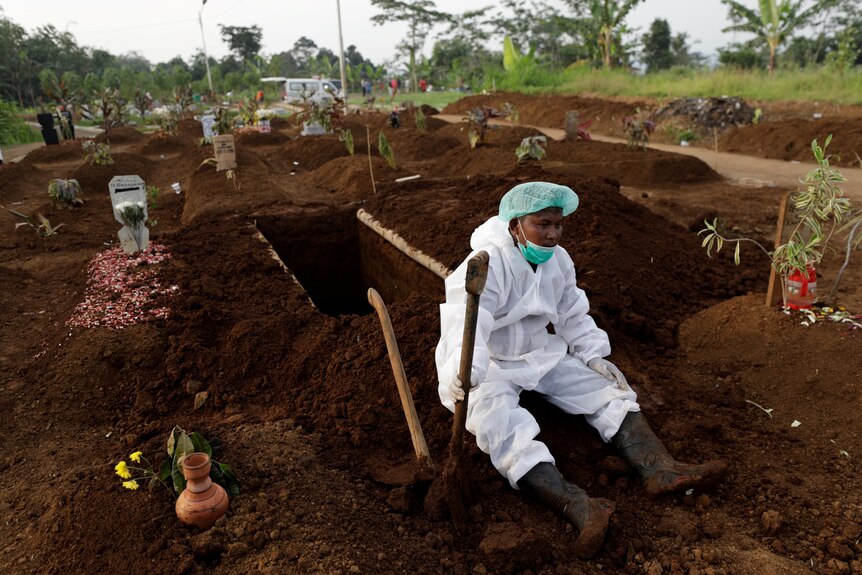 A man in full PPE sits down holding a shovel in front of an open grave 
