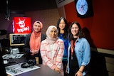 Four girls in a radio studio look at the camera smiling. A red box with 'triple j' and a drum in white writing is lit up.