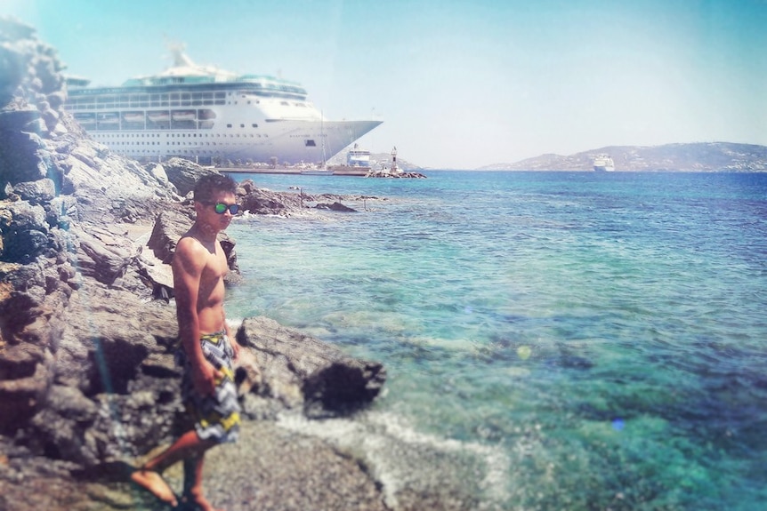 A young man in board shorts on a rocky shore with a cruise ship behind him