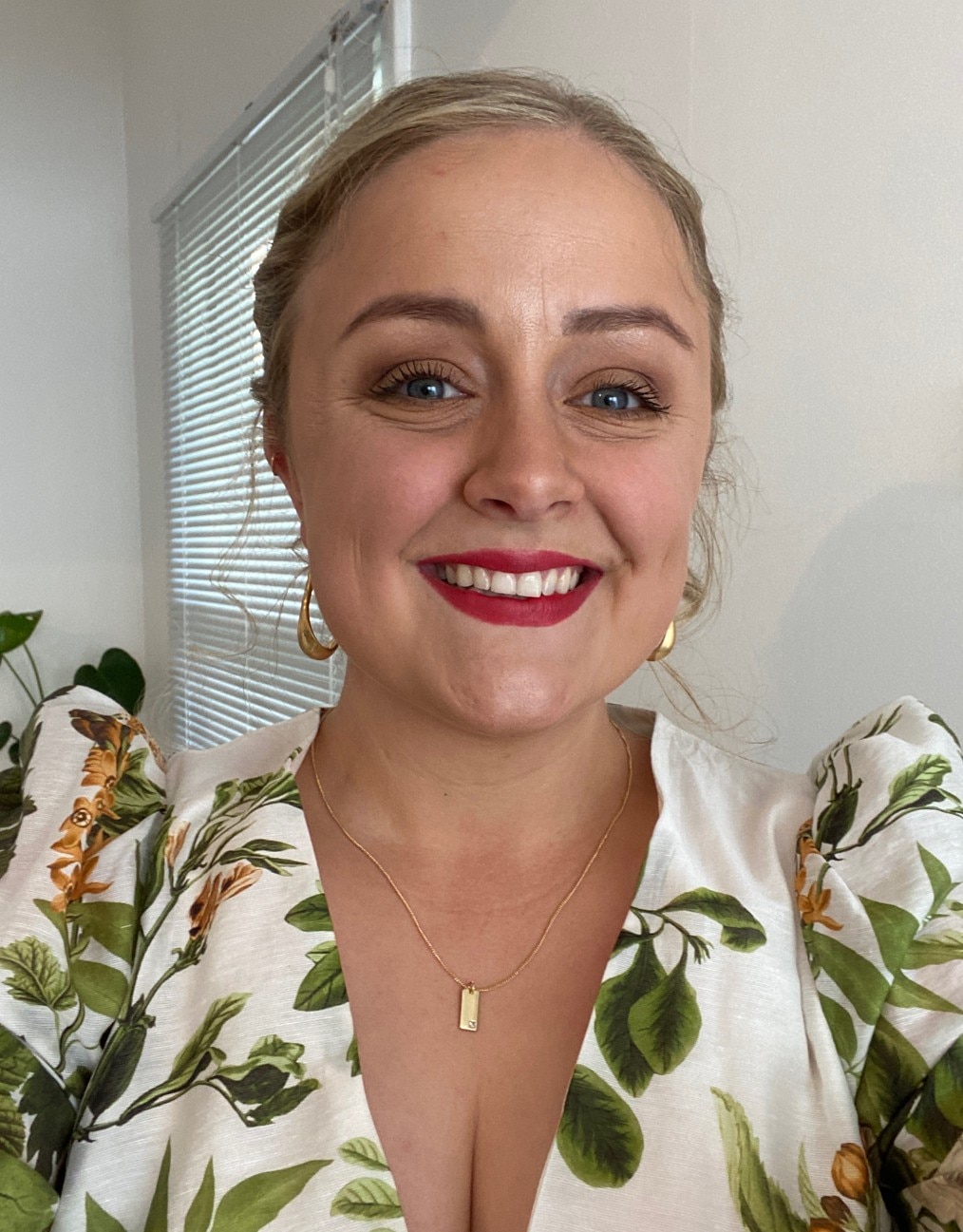 A woman with tied back blonde hair smiles wide for a selfie. She wears a green and white dress and red lipstick.