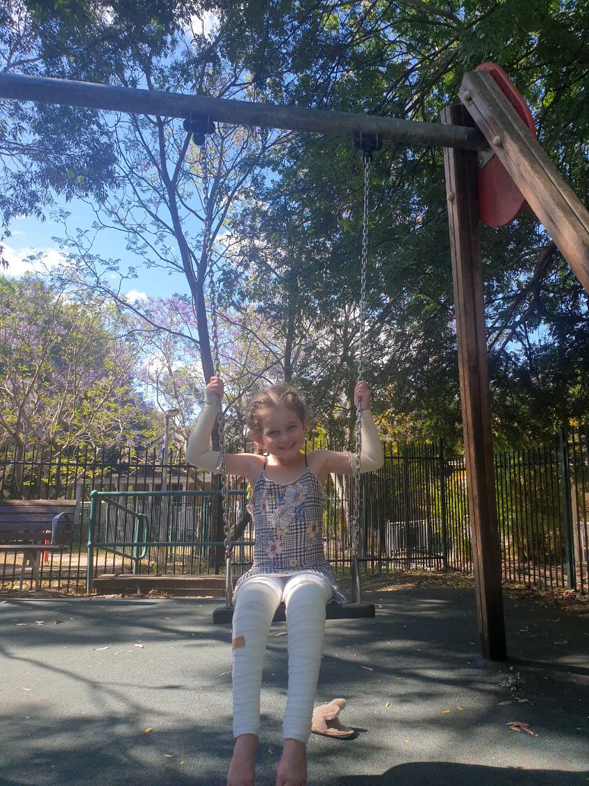 Brunette young girl sitting on swing with white bandaged limbs smiling
