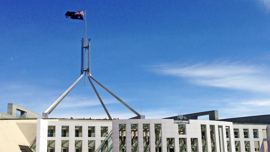 People stand outside the front entrance to Parliament House in Canberra. The sky is blue with a few white clouds.