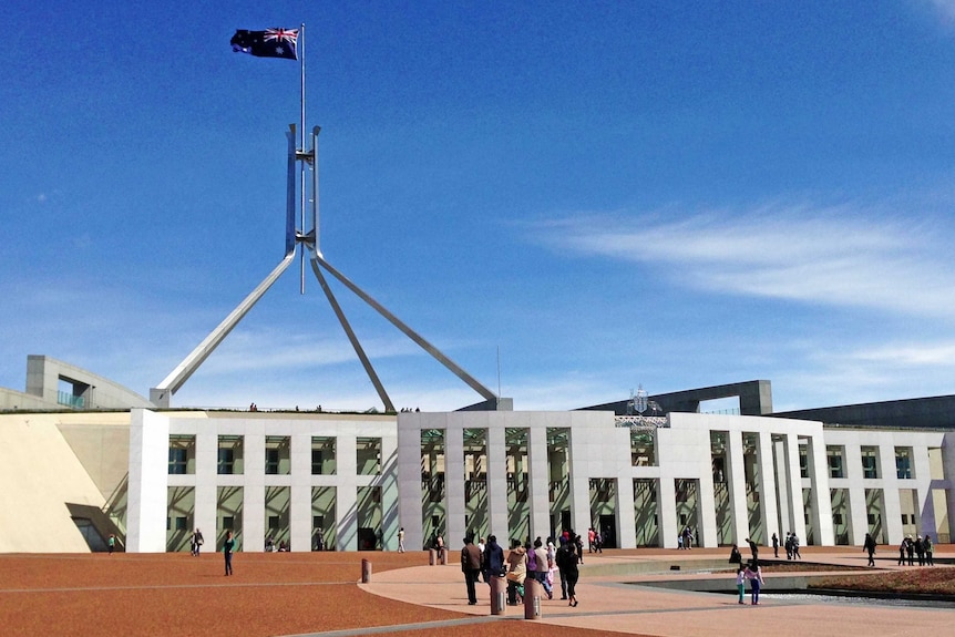 People stand outside the front entrance to Parliament House in Canberra. The sky is blue with a few white clouds.