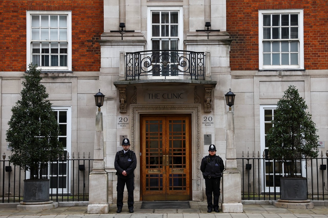 Two police officers guard the door of a large, grand building seen from across a street.