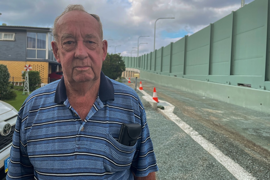 An elderly man standing in front of brick homes with green highway sound barriers in the background.