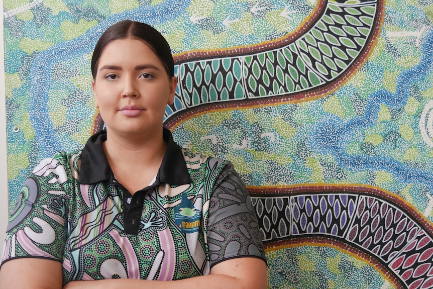 A woman with dark hair stands in front of an Aboriginal painting. She is wearing a polo shirt.