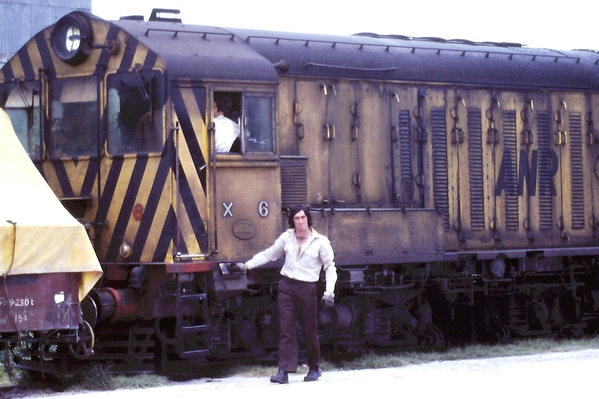 A man in 1970s collared shirt stands beside a diesel locomotive.