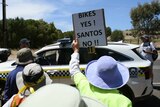 A crowd of people around a police car, one is holding a placard that says 'Bikes yes Santos no'
