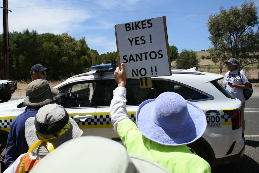 A crowd of people around a police car, one is holding a placard that says 'Bikes yes Santos no'