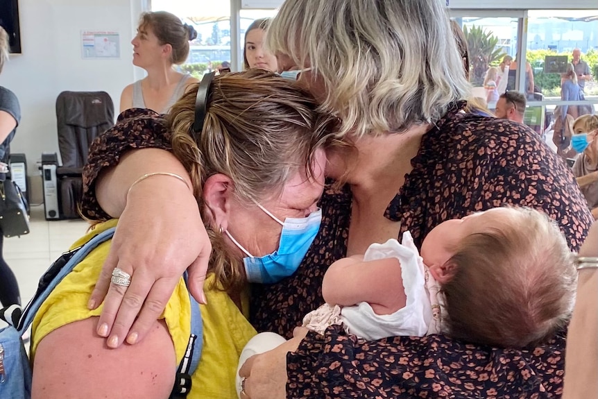 Sisters Diane Senior and Lesley Eva hug with a baby at the airport. 