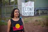 A photo of woman wearing a shirt with Indigenous flag on the chest with trees and protest sign in the background.