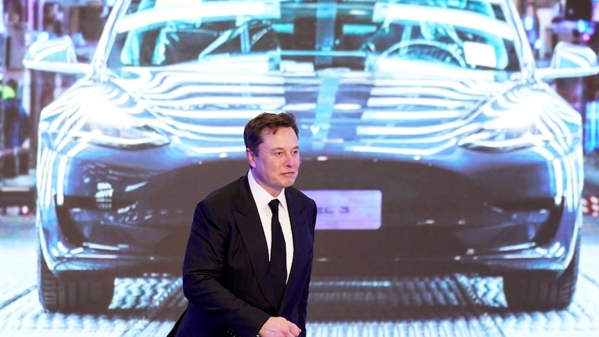 A man in a dark suit and white shirt walks in front of an illuminated dark-coloured car.