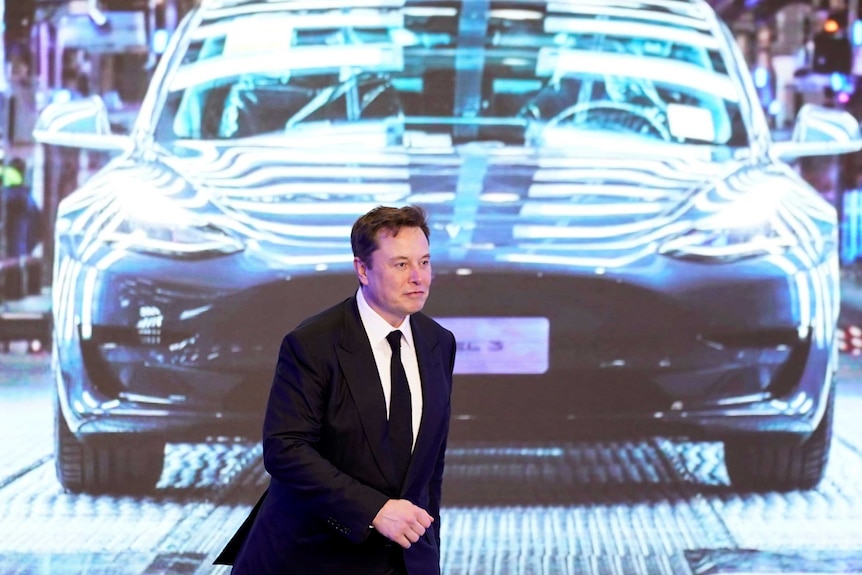 A man in a dark suit and white shirt walks in front of an illuminated dark-coloured car.