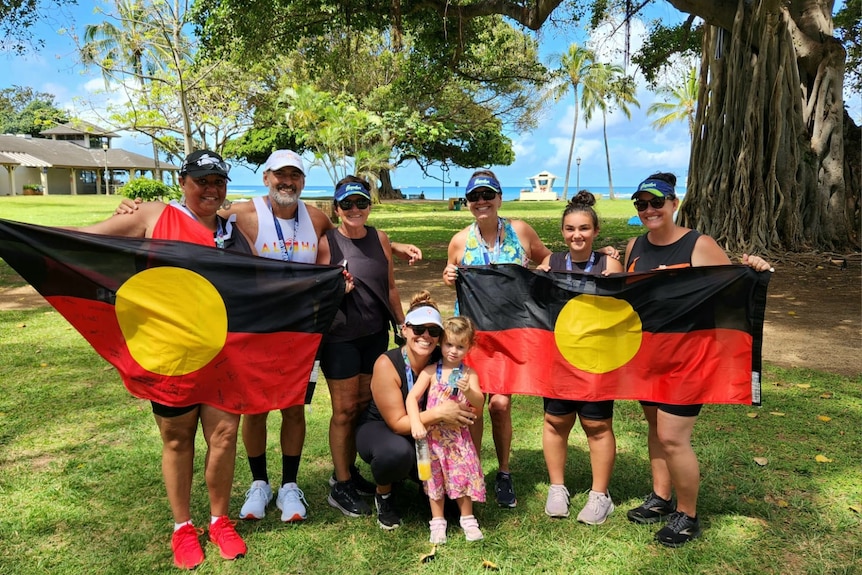 A group of runners in their middle age standingn underneath a large fig tree, smiling, with two Aboriginal flags unfurled.