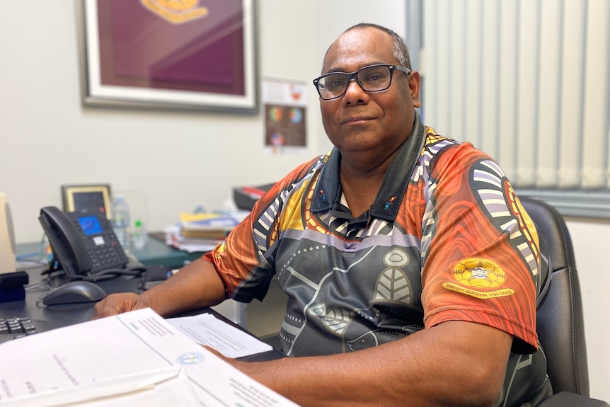An man in a shirt featuring Indigenous designs sits at a desk.