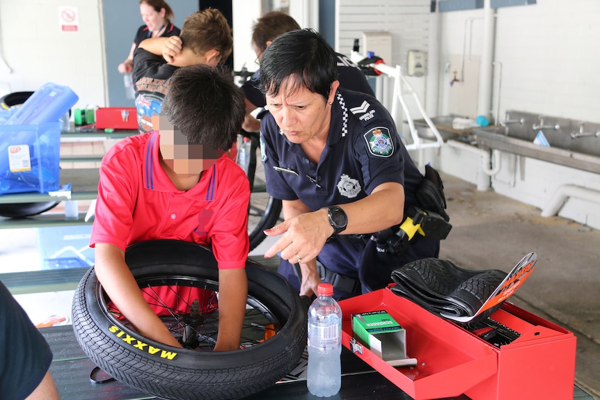 A female police officer guiding a boy repairing a bicycle wheel