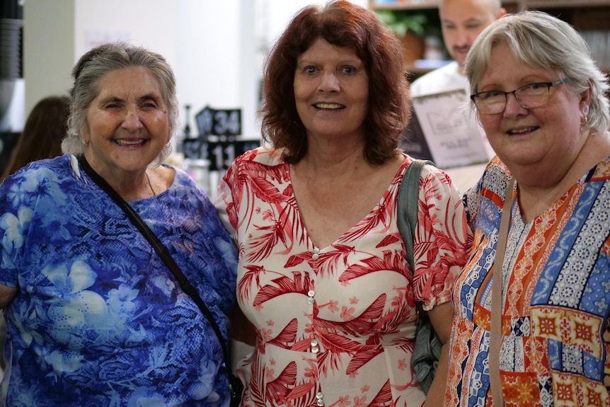 Three older women stand together wearing printed shirts, they are in front of a cafe front.