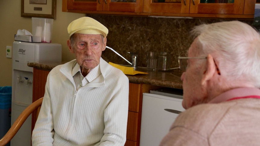 An elderly man wearing a yellow cap sitting at a table talking to another man.