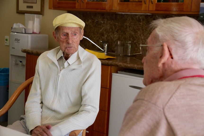 An elderly man wearing a yellow cap sitting at a table talking to another man.
