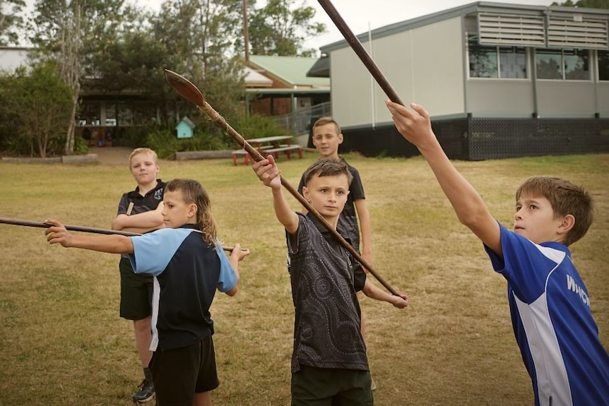 Three boys on school grounds in uniform holding up long spears about to throw them, two boys behind watching 