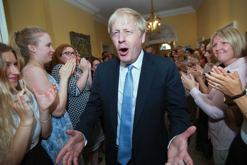 Boris Johnson pulls a gleeful face with his hands wide as he walks through a corridor surrounding by clapping staff.