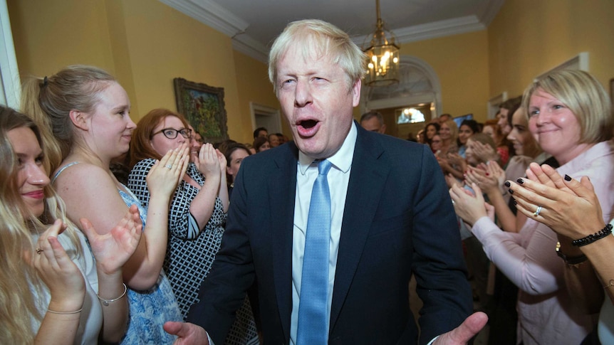 Boris Johnson pulls a gleeful face with his hands wide as he walks through a corridor surrounding by clapping staff.