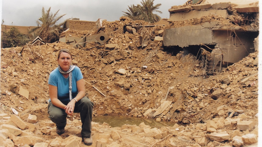 A woman with a concerned face squats next to a hole in the ground where a house had stood, caused by aerial bombardment