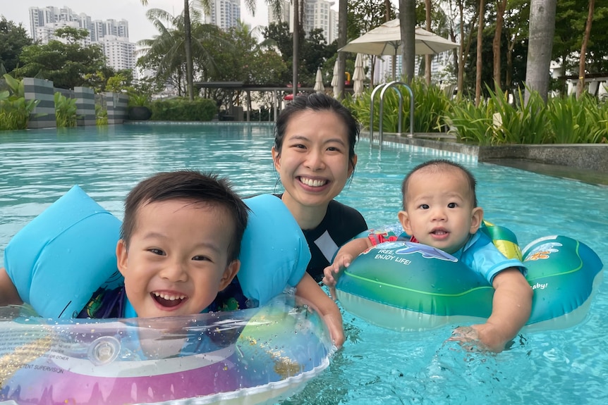 A 30-something Chinese lady stands and smiles in swimming pool along two floating toddlers in tropical setting.