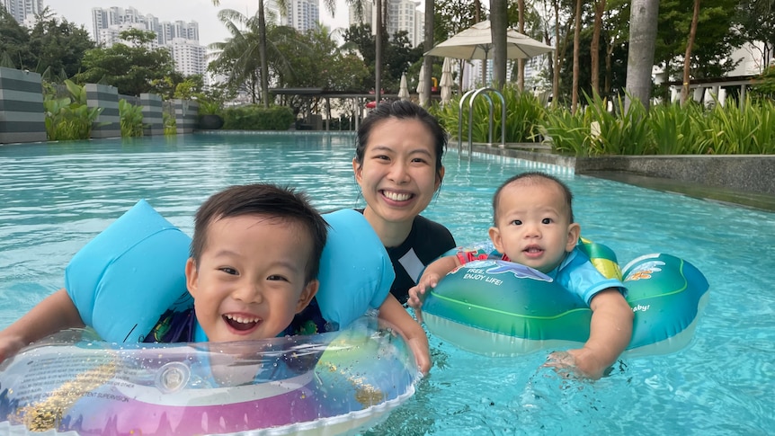 Una donna cinese di 30 anni in piedi e sorridente in piscina con due bambini piccoli che galleggiano in un ambiente tropicale.