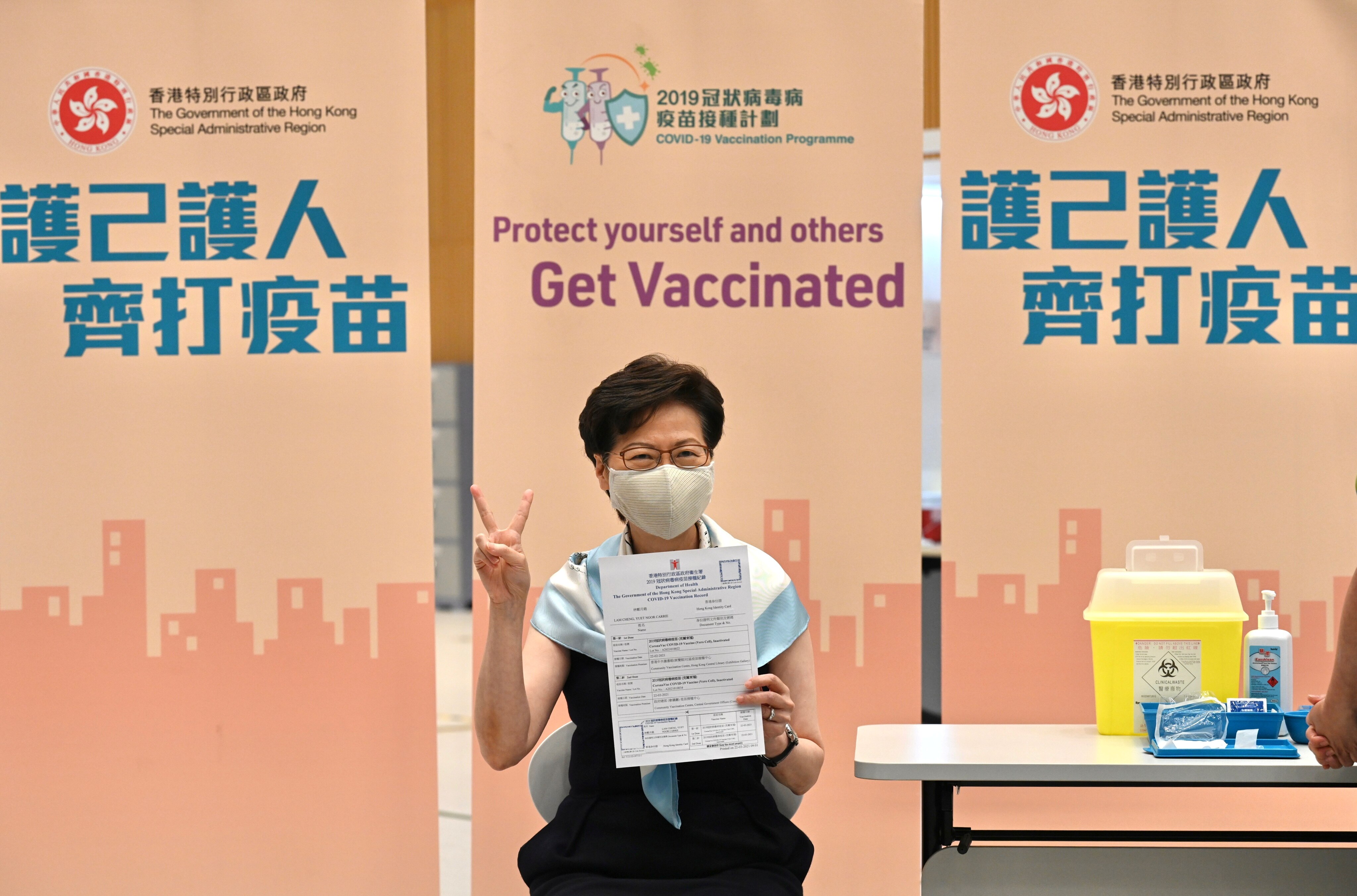 A middle-aged Asian woman in face mask gives a peace sign and holds up a white certificate in a medical clinic.