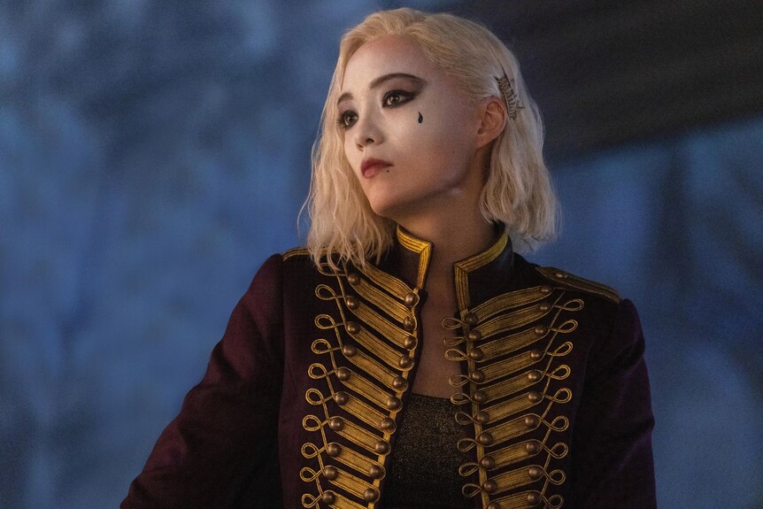 Pom Klementieff, a white woman with bleach blonde hair and dark eyes, wears a red and gold rococo jacket and white face paint.