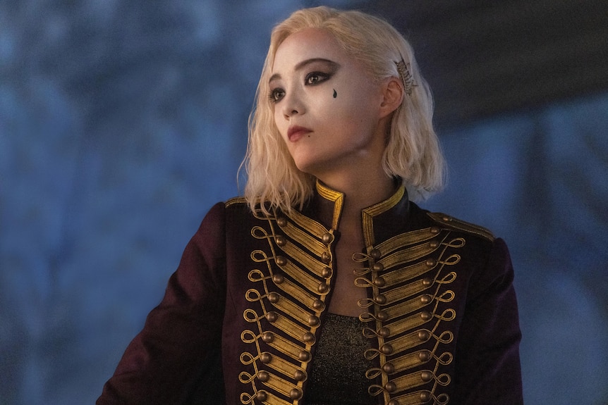 Pom Klementieff, a white woman with bleach blonde hair and dark eyes, wears a red and gold rococo jacket and white face paint.