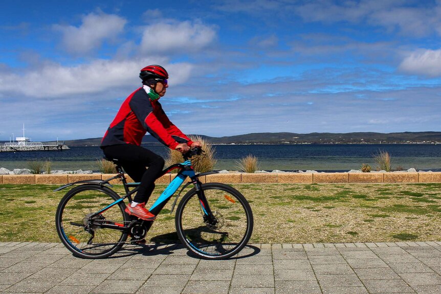 A man rides an electric bicycle across the frame with a harbour and blue sky in the background.