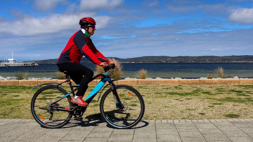 A man rides an electrical bicycle across the frame with a harbour and blue sky in the background.