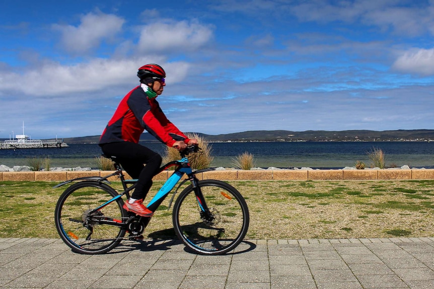 A man rides an electric bicycle across the frame with a harbour and blue sky in the background.