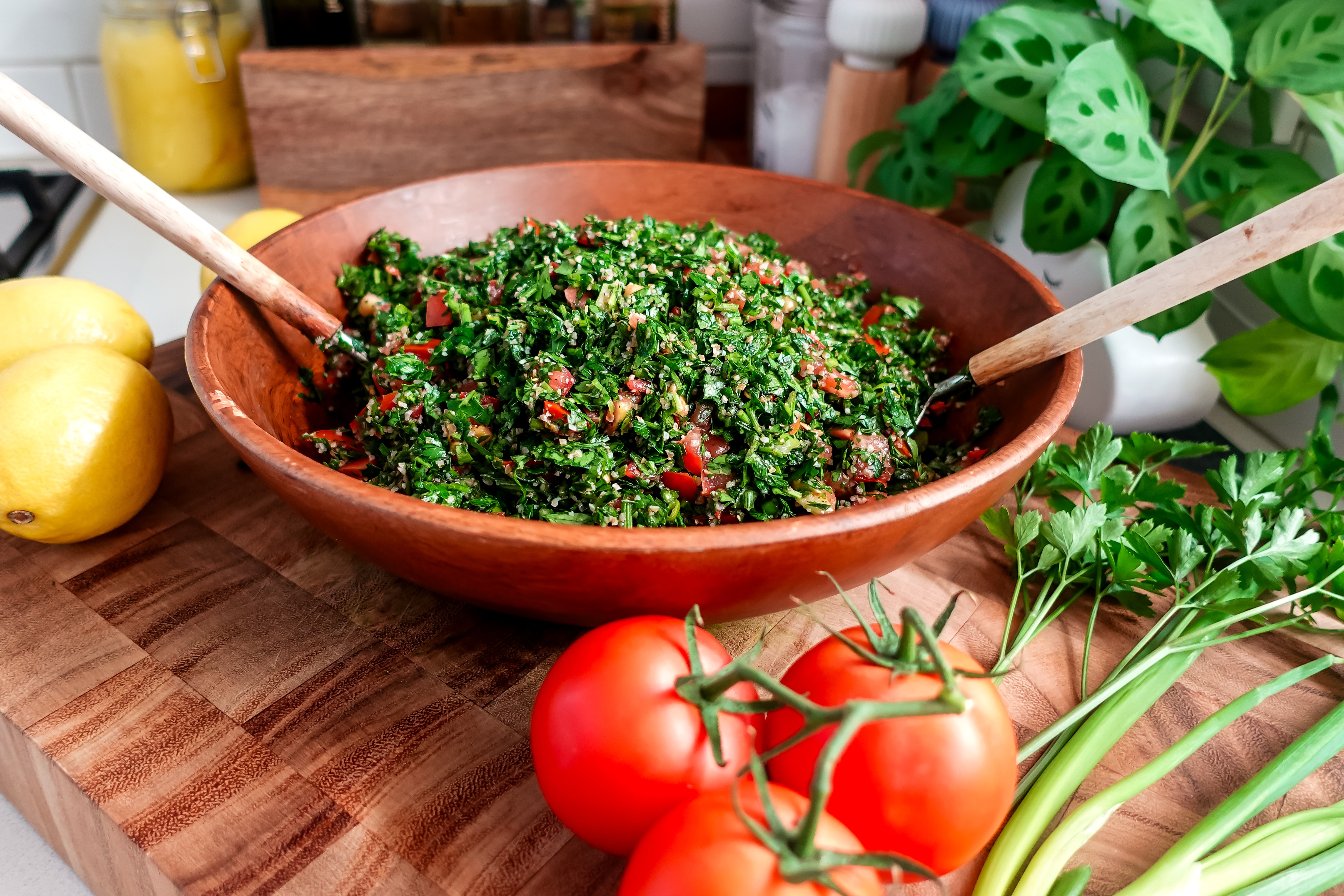A wooden salad bowl with tabouli and salad servers. Tomatoes, spring onions and herbs nearby.