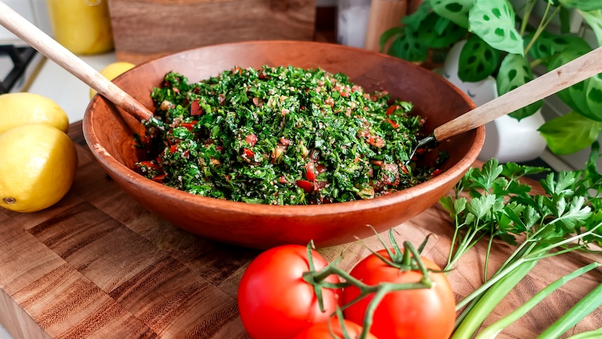 A wooden salad bowl with tabouli and salad servers. Tomatoes, spring onions and herbs nearby.