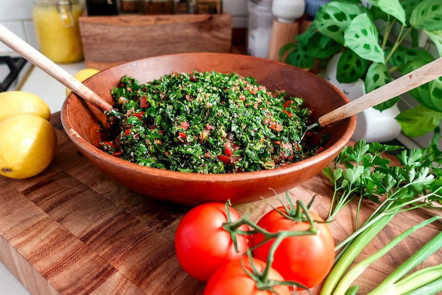 A wooden salad bowl with tabouli and salad servers. Tomatoes, spring onions and herbs nearby.