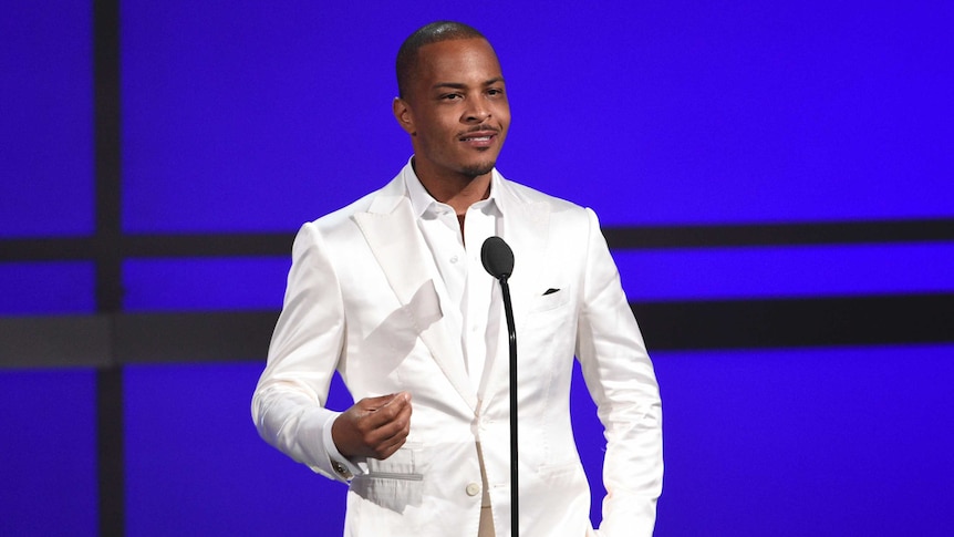 T.I. stands on stage in front of a microphone. He's wearing a white suit.