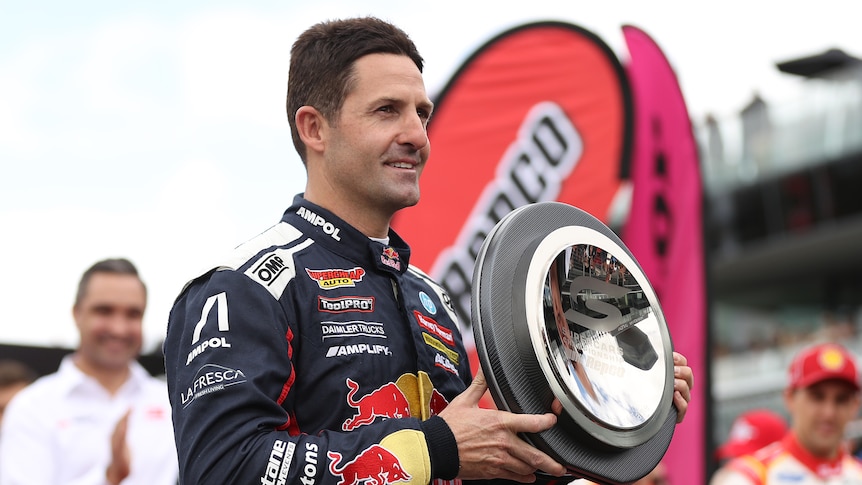 A supercars driver stands holding a trophy in the shape of a car racing tyre honouring him before his final race.
